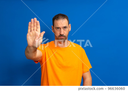 Bearded hispanic man dressed in orange t-shirt making stop gesture with palm of hand isolated on blue studio background. He is fed up with the social and labor injustices caused by the current system. Bearded hispanic man dressed in orange t-shirt making stop gesture with palm of hand isolated on blue studio background. He is fed up with the social and labor injustices caused by the current system. 96636060