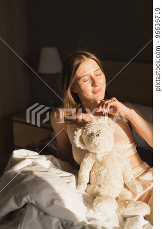 Portrait of girl is waking up in morning stretches in bed and sun shines from window. Happy young woman greets new sunny day and holds teddy bear toy Portrait of girl is waking up in morning stretches in bed and sun shines from window. Happy young woman greets new sunny day and holds teddy bear toy 96636719