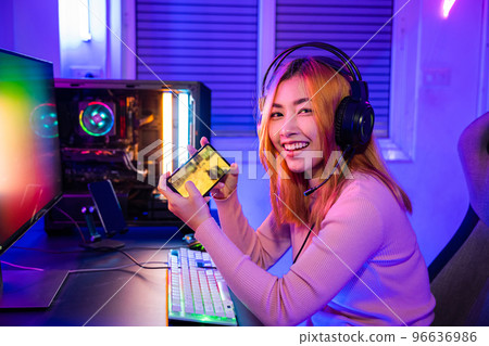Gamer playing online game application on mobile phone wear gaming headphones looking to camera, Smiling young woman live stream she play video game via smartphone at home neon lights living room Gamer playing online game application on mobile phone wear gaming headphones looking to camera, Smiling young woman live stream she play video game via smartphone at home neon lights living room 96636986