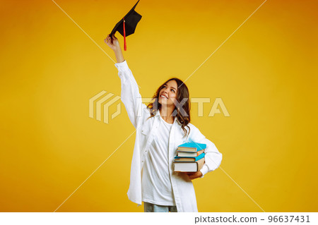 Graduate woman in a graduation hat on her head, with books stands on a yellow background. 96637431