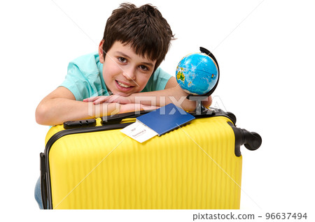 Close-up portrait of a happy teenage boy passenger with yellow suitcase and globe, cutely smiles looking at camera, isolated over white background. Travel. Tourism. Journey concept Free ad space 96637494