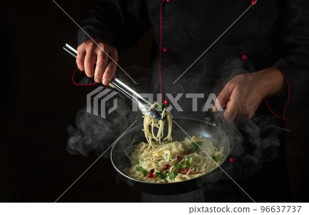The chef cooks delicious spaghetti with spices and seasonings in a steamed hot pan. Space for advertising on a dark background. Menu for a restaurant or hotel. The chef cooks delicious spaghetti with spices and seasonings in a steamed hot pan. Space for advertising on a dark background. Menu for a restaurant or hotel. 96637737