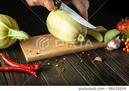 Slicing zucchini with a knife before cooking by chef's hands on a wooden cutting board. The thick flesh of a vegetable marrow is used in the UK. Slicing zucchini with a knife before cooking by chef's hands on a wooden cutting board. The thick flesh of a vegetable marrow is used in the UK. 96638834