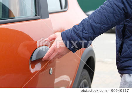 Hand on handle. Man in blue jacket opens door of his orange car in parking lot. Close up. Hand on handle. Man in blue jacket opens door of his orange car in parking lot. Close up. 96640517