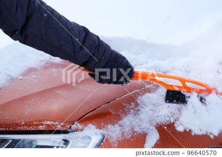 Snowy. Brush in mans hand. Man clears orange car from snow. Car covered with snow. Winter problems of car drivers. Snowy. Brush in mans hand. Man clears orange car from snow. Car covered with snow. Winter problems of car drivers. 96640570