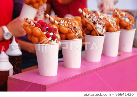 Bubble waffles with raspberries, chocolate and marshmallow in white paper cups are sold on Open kitchen food festival event. Street food in food stall. Bubble waffles with raspberries, chocolate and marshmallow in white paper cups are sold on Open kitchen food festival event. Street food in food stall. 96641420