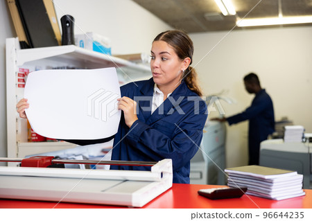 Attentive young female in a blue uniform verifying printed sheet during work in the printer shop 96644235