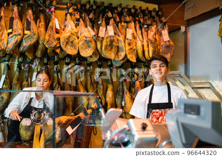 Young female and male vendors arranging jamon at counter in butcher shop 96644320