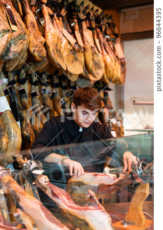 Young salesman in black uniform slicing jamon at counter in butcher shop Young salesman in black uniform slicing jamon at counter in butcher shop 96644395