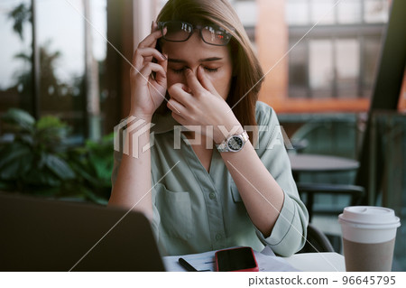 A tired and stressed young woman holds glasses and massages his nose while working hard in a cafe. 96645795