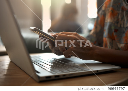 Close-up of woman's hand using smartphone while working on laptop in a coffee shop. Close-up of woman's hand using smartphone while working on laptop in a coffee shop. 96645797
