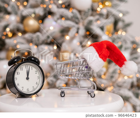Christmas gifts shopping time. A shopping trolley with a santa hat and an alarm clock by the Christmas tree. Christmas gifts shopping time. A shopping trolley with a santa hat and an alarm clock by the Christmas tree. 96646423