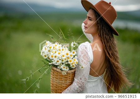 A middle-aged woman in a white dress and brown hat stands on a green field and holds a basket in her hands with a large bouquet of daisies. In the background there are mountains and a lake. 96651915