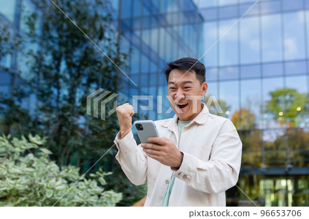 Asian businessman in casual clothes with phone outside office building celebrating victory and success, man joyfully holding hand up triumph gesture. Asian businessman in casual clothes with phone outside office building celebrating victory and success, man joyfully holding hand up triumph gesture. 96653706
