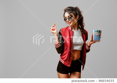 Portrait of pretty stylish brunette girl in sunglasses holding pop corn bowl over gray background 96654035