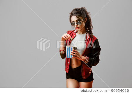 Portrait of beautiful stylish brunette girl in sunglasses holding pop corn bowl on gray background 96654036