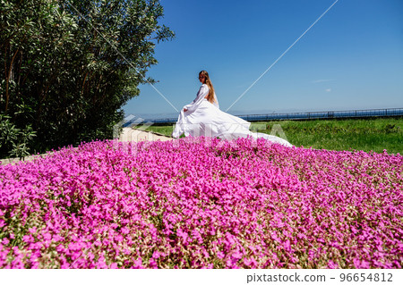 a beautiful woman in a white flowing long dress stay near a beautiful field with pink flowers rear view 96654812