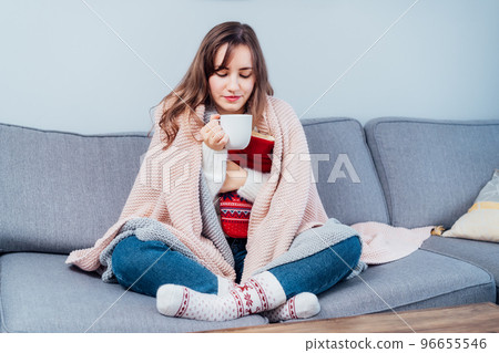 Woman freezes in wintertime. Young girl wearing warm woolen socks and wrapped into two blankets, holding a cup of hot drink and heating pad while sitting on sofa at home. Keep warm. Selective focus Woman freezes in wintertime. Young girl wearing warm woolen socks and wrapped into two blankets, holding a cup of hot drink and heating pad while sitting on sofa at home. Keep warm. Selective focus 96655546