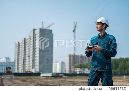 A man in a helmet and overalls controls a drone at a construction site. The builder carries out technical oversight. 96655785