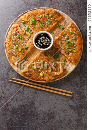 Kimchijeon South Korean pancake incorporates sliced kimchi served with a dipping sauce and sprinkled with sesame seeds and green onion closeup on the wooden board. Vertical top view 96658150