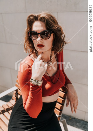 Portrait of a woman on the street. An attractive woman in glasses, a red blouse and a black skirt is sitting on a bench outside. 96659683