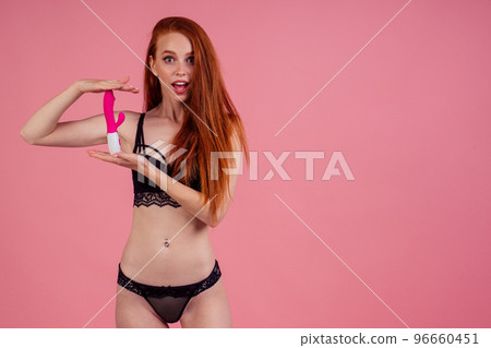 sex education concept.beautiful amazed and surprised redhaired ginger woman in sexy black linen lace holding vibro dildo in studio pink background 96660451