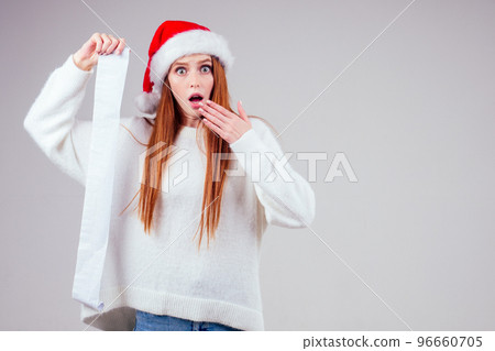amazed and surprised redhaired ginger woman wearing red santa claus hat and knitted sweater holding long shopping receipt list in studio white background. big money xmas spending 96660705