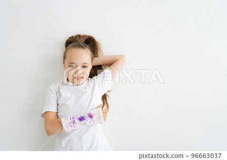 High-angle view of positive sweet little girl with broken hand wrapped in plaster bandage with colorful draw lying on white floor in studio looking at camera. Concept of child insurance and healthcare 96663037