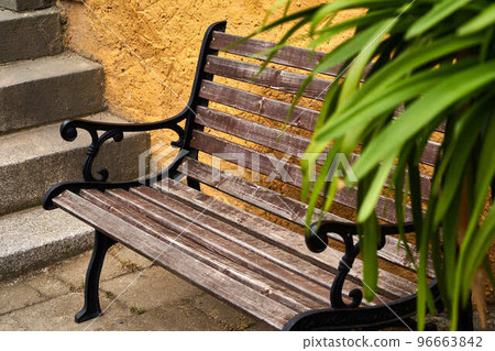 Brown weathered bench with cast iron sides behind a stone staircase in front of a blurred green plant, selective focus Brown weathered bench with cast iron sides behind a stone staircase in front of a blurred green plant, selective focus 96663842