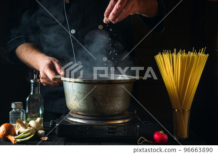 The chef prepares Italian pasta on the background of vegetables. Close-up of a cook hand pouring peppers into saucepan in a restaurant kitchen 96663890