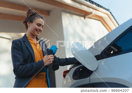Young woman holding power supply cable from her electric car, prepared for charging it in home, sustainable and economic transportation concept. Young woman holding power supply cable from her electric car, prepared for charging it in home, sustainable and economic transportation concept. 96664207
