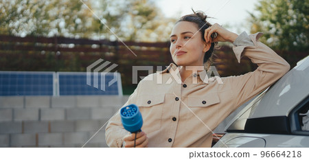Young woman holding power supply cable from her electric car, prepared for charging it in home, sustainable and economic transportation concept. 96664218