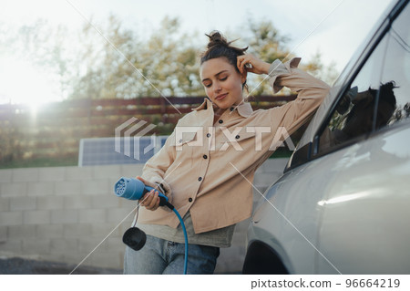 Young woman holding power supply cable from her electric car, prepared for charging it in home, sustainable and economic transportation concept. 96664219