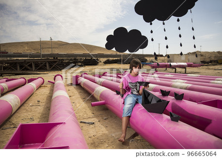a cute kid in a pink t-shirt with a cat print plays among pink pipes with black clouds, rain, boats and paper planes against the blue sky in an industrial location. fashion style photoshoot a cute kid in a pink t-shirt with a cat print plays among pink pipes with black clouds, rain, boats and paper planes against the blue sky in an industrial location. fashion style photoshoot 96665064