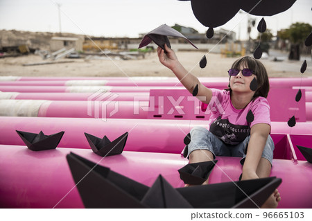 a cute kid in a pink t-shirt with a cat print plays among pink pipes with black clouds, rain, boats and paper planes against the blue sky in an industrial location. fashion style photoshoot 96665103