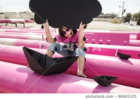a cute kid in a pink t-shirt with a cat print plays among pink pipes with black clouds, rain, boats and paper planes against the blue sky in an industrial location. fashion style photoshoot a cute kid in a pink t-shirt with a cat print plays among pink pipes with black clouds, rain, boats and paper planes against the blue sky in an industrial location. fashion style photoshoot 96665105