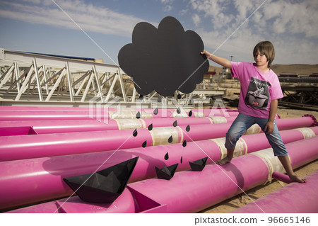 a cute kid in a pink t-shirt with a cat print plays among pink pipes with black clouds, rain, boats and paper planes against the blue sky in an industrial location. fashion style photoshoot a cute kid in a pink t-shirt with a cat print plays among pink pipes with black clouds, rain, boats and paper planes against the blue sky in an industrial location. fashion style photoshoot 96665146