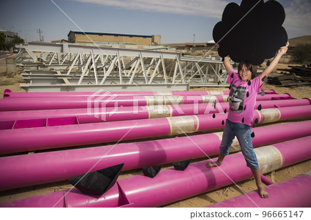 a cute kid in a pink t-shirt with a cat print plays among pink pipes with black clouds, rain, boats and paper planes against the blue sky in an industrial location. fashion style photoshoot a cute kid in a pink t-shirt with a cat print plays among pink pipes with black clouds, rain, boats and paper planes against the blue sky in an industrial location. fashion style photoshoot 96665147