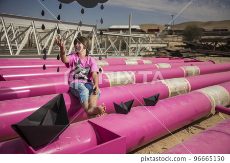 a cute kid in a pink t-shirt with a cat print plays among pink pipes with black clouds, rain, boats and paper planes against the blue sky in an industrial location. fashion style photoshoot a cute kid in a pink t-shirt with a cat print plays among pink pipes with black clouds, rain, boats and paper planes against the blue sky in an industrial location. fashion style photoshoot 96665150