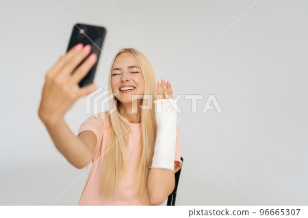 Studio portrait of positive young blonde woman with broken arm wrapped in plaster bandage talking smartphone making video call, taking selfie picture, standing on white isolated background. Studio portrait of positive young blonde woman with broken arm wrapped in plaster bandage talking smartphone making video call, taking selfie picture, standing on white isolated background. 96665307