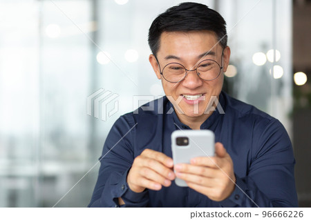 Close-up photo of a happy and smiling Asian man reading happy news from the phone, a businessman in a shirt and glasses smiling and rejoicing, a man works inside the office near the window. Close-up photo of a happy and smiling Asian man reading happy news from the phone, a businessman in a shirt and glasses smiling and rejoicing, a man works inside the office near the window. 96666226