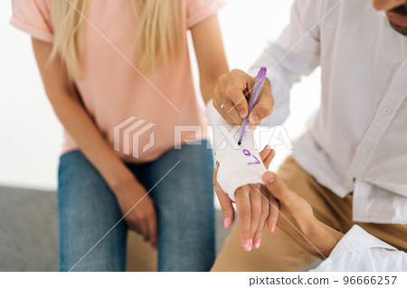 Close-up high-angle view of unrecognizable male friend writing joking inscription on cast of woman with broken hand wrapped in plaster bandage. Concept of healthcare, friendship and fun. 96666257