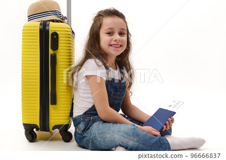 Adorable little girl passenger wearing denim overalls, sitting over white background with boarding pass in her hands, leaning on yellow suitcase and smiling a cheerful toothy smile, looking at camera Adorable little girl passenger wearing denim overalls, sitting over white background with boarding pass in her hands, leaning on yellow suitcase and smiling a cheerful toothy smile, looking at camera 96666837