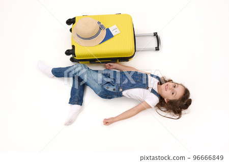 Top view cute little girl tourist in denim overalls, with suitcase, boarding pass and straw hat, lying on white background, advertising space. Travel abroad. getaway. Air flight trip journey concept Top view cute little girl tourist in denim overalls, with suitcase, boarding pass and straw hat, lying on white background, advertising space. Travel abroad. getaway. Air flight trip journey concept 96666849