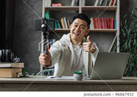Attractive asian man in casual clothes taking selfie on his smart phone using stick showing thumb up Attractive asian man in casual clothes taking selfie on his smart phone using stick showing thumb up 96667452