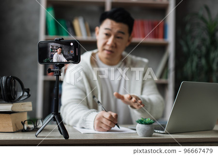 Focus on screen, smiling asian man talking and gesturing in front of modern smartphone camera Focus on screen, smiling asian man talking and gesturing in front of modern smartphone camera 96667479