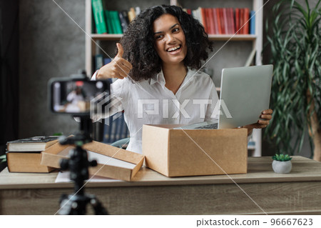 Multicultural woman recording video on phone camera while unpacking box with new wireless laptop. 96667623