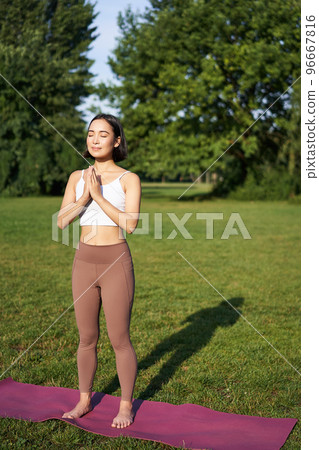 Vertical shot of asian woman standing in asana, doing yoga exercises on fresh air in park, wearing leggings, standing on rubber mat 96667816