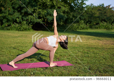Vertical shot of young korean woman doing yoga training on rubber mat, making asana exercises on fresh air in park Vertical shot of young korean woman doing yoga training on rubber mat, making asana exercises on fresh air in park 96668011