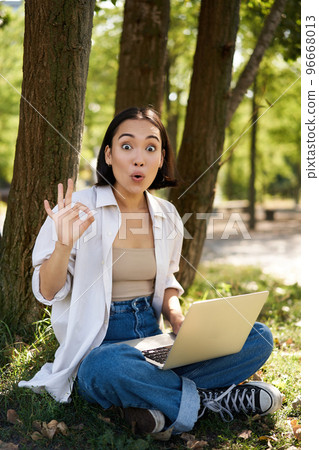 Portrait of asian young woman, student doing homework, working in park, sitting beside tree with laptop and showing okay sign, approve smth 96668013